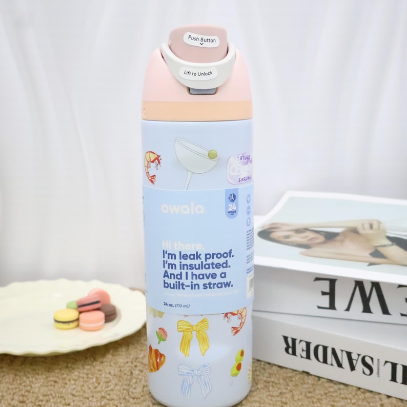May include: A pastel-colored Owala water bottle with a push-button lid. The bottle features a light blue section with food-themed stickers and text that reads, "Hi there. I'm leak proof. I'm insulated. And I have a built-in straw." It holds 34 oz.