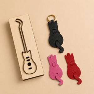 May include: A rectangular wooden board with a metal outline of a guitar shape, alongside three cat-shaped keychains in black, pink, and red. The black keychain features a gold-toned metal ring at the top, while all three have a tail-like extension.