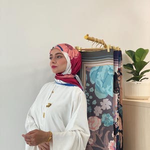 Op de afbeelding: Een vrouw draagt een witte jurk en een hoofddoek met bloemenpatroon. De hoofddoek heeft roze, rode en blauwe bloemenmotieven. Een goudkleurige broche en armband zijn zichtbaar. Verschillende stoffen met patronen hangen op de achtergrond.