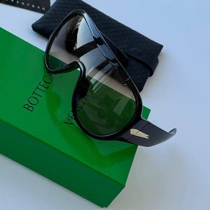 Könnte beinhalten: Schwarze Sonnenbrille im Shield-Design mit einem durchgehenden Glas und goldfarbenen Metallakzenten an den Bügeln. Die Sonnenbrille wird mit einem schwarzen, strukturierten Schutzetui und einer grünen Box mit dem Aufdruck Bottega Veneta geliefert.