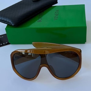 May include: Brown-tinted sunglasses with a thick, rounded frame and dark lenses, positioned in front of a bright green Bottega Veneta box and a black, woven-textured protective case. The brand name is visible on the lens and the box.