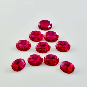 May include: A collection of nine oval-shaped, faceted red gemstones arranged in a pyramid formation. Each stone features a brilliant cut with multiple reflective surfaces that catch the light, displaying a deep, vibrant crimson colour throughout.