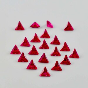 Puede incluir: Una colección de veinte pequeños cristales triangulares de color fucsia vibrante. Cada pieza presenta una superficie facetada que refleja la luz, creando una forma geométrica afilada con una parte superior puntiaguda y una base plana.