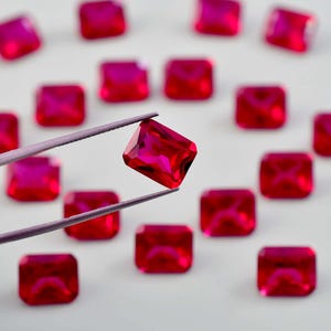 May include: A collection of rectangular, faceted, vibrant red gemstones scattered across a surface. One individual red gemstone is held by a pair of metal tweezers, showcasing its clear, polished surface and sharp, geometric edges.