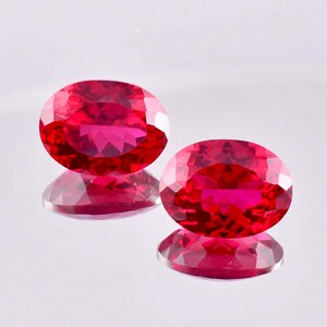 Puede incluir: Dos piedras preciosas de color rojo vibrante con corte ovalado y una superficie facetada que refleja la luz. Las piedras están colocadas una al lado de la otra, mostrando un tono carmesí profundo y brillante con un aspecto pulido.