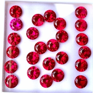 Puede incluir: Una colección de veinte piedras preciosas redondas, facetadas y de color rojo vibrante dispuestas en un patrón de cuadrícula. Cada piedra presenta un corte brillante con múltiples facetas triangulares que reflejan la luz y brillan.