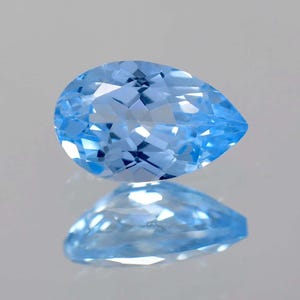 Puede incluir: Una gema facetada en forma de pera, de un vibrante tono azul claro. La superficie de la gema refleja la luz, creando un efecto brillante. La piedra se muestra sobre una superficie reflectante, con su imagen reflejada debajo.