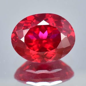 May include: An oval-cut gemstone with a vibrant, deep red colour and a faceted surface that creates multiple light reflections. The stone displays a clear, transparent quality with sharp geometric edges and a polished finish throughout its structure.
