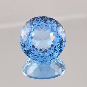 May include: A faceted, round, light blue gemstone. The gem is highly reflective, with a complex pattern of internal facets that create a sparkling effect. The gem is sitting on a reflective surface, creating a mirrored image.