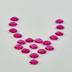 Op de afbeelding: Een verzameling van vijftien levendige fuchsia roze marquise-geslepen edelstenen gerangschikt in een V-vormig patroon. Elke individuele steen heeft een gefacetteerd oppervlak dat licht reflecteert, wat zorgt voor een schitterende uitstraling.