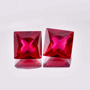 Puede incluir: Dos piedras preciosas de color rojo vibrante con corte cuadrado y superficies facetadas que crean un patrón geométrico. Las piedras son transparentes y muestran un tono carmesí profundo, reflejando la luz en sus múltiples caras.