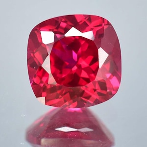 Puede incluir: Una piedra preciosa de corte cojín con un color rojo vibrante y saturado, y una superficie facetada que crea reflejos de luz geométricos nítidos. La piedra muestra gran claridad y brillo, con un reflejo distintivo visible justo debajo.