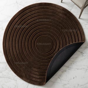 May include: A circular dark brown rug featuring a series of concentric embossed rings across its surface. The text FZRUGSART is visible multiple times across the rug. One corner of the rug is folded over to reveal a solid black backing material.