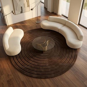 May include: A large circular dark brown rug with concentric ring patterns, featuring two cream-colored curved sofas and a round glass coffee table with a sculptural base. The rug displays the text FZRUGSART in multiple locations.