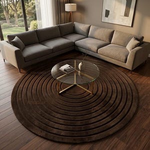 May include: A large, circular, dark brown area rug featuring a concentric ring pattern with deep, carved-style grooves. The rug is positioned on a dark wood floor beneath a glass-topped coffee table with a gold-toned metal base.