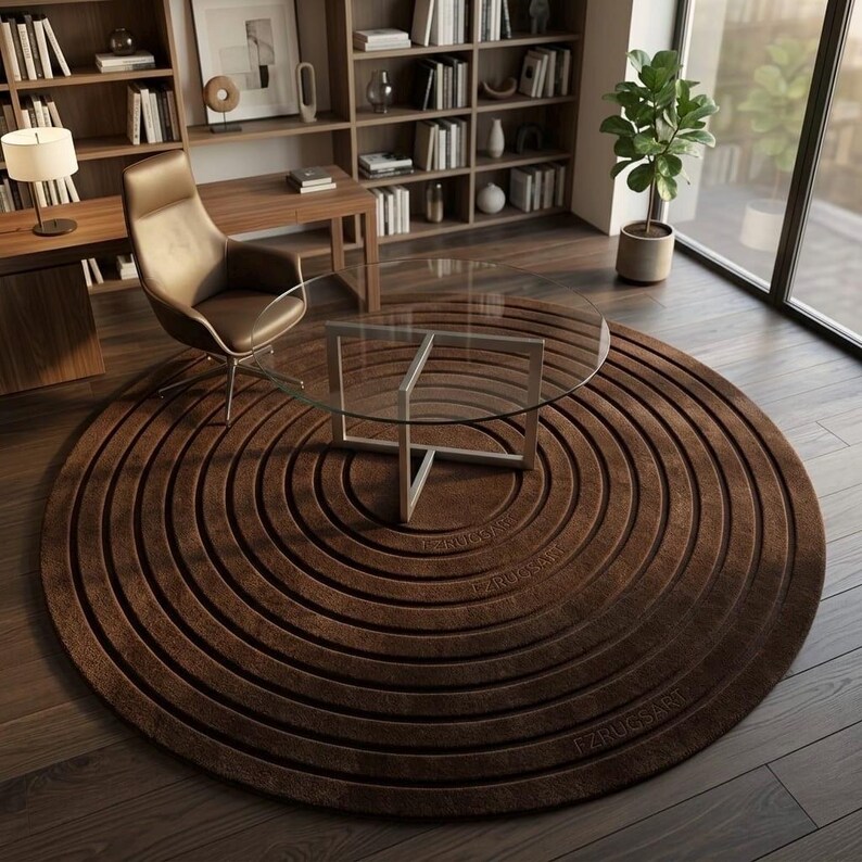 May include: A large, circular, dark brown rug featuring a concentric ring pattern with the text FZRUGSART embossed on the surface. The rug is positioned under a round glass-topped table with a silver metal base, resting on a dark wood floor.