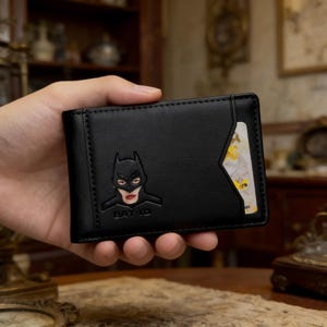 May include: Black leather wallet featuring a Batman-inspired design. The wallet has a card slot and a debossed image of a female Batman character with red lipstick. The text "BAT ID" is below the image.