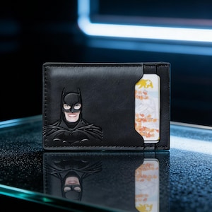 May include: Black leather wallet featuring an embossed Batman design on the front. The wallet has a card slot visible on the right side. The image is set against a dark background with a neon blue light.