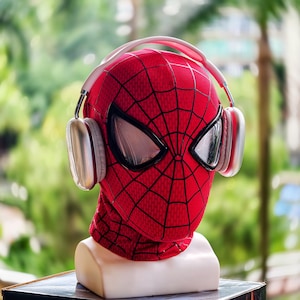 May include: A red Spider-Man mask with a black web pattern and white eye lenses, paired with white and silver headphones. The mask is displayed on a white bust, set against a blurred green background.