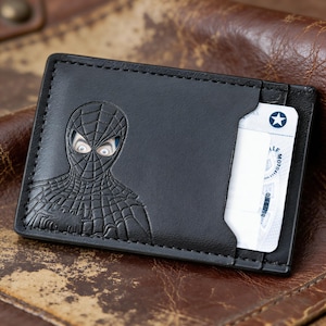 May include: Black leather wallet featuring an embossed Spider-Man design. The wallet has a card slot with a white card partially visible. The wallet is rectangular and has a stitched border.