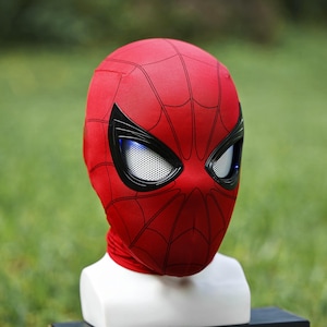 May include: A red fabric mask featuring a black web pattern across the surface. The mask includes large, angular black-rimmed eye lenses with white mesh detailing and integrated blue LED lighting elements visible within the eye openings.