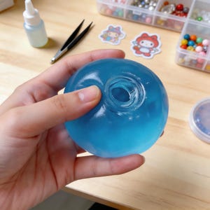 Pode incluir: Uma esfera azul translúcida com um buraco central é segurada. Materiais de artesanato, incluindo uma garrafa pequena, pinças e adesivos, estão por perto. A esfera parece ser um material de artesanato.