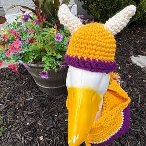 May include: A crochet hat featuring two white horns, a yellow crown, and a purple brim, worn by a white goose statue with a yellow beak. The goose is also wearing a yellow and purple crochet garment that covers its neck and chest area.