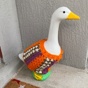 May include: A white goose figurine wearing a crocheted sweater with orange, brown, purple, and white stripes. The goose is standing on a green base with a yellow beak and black eyes. The goose is standing on a brown doormat with white stars.