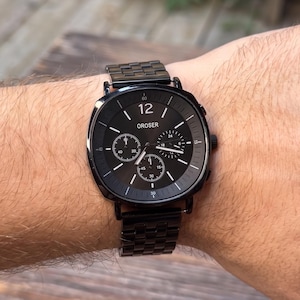 Puede incluir: Un reloj de pulsera negro con una esfera cuadrada y una correa de metal negro a juego. La esfera del reloj muestra la marca "OROSER" y presenta tres subesferas. El reloj se lleva en la muñeca.