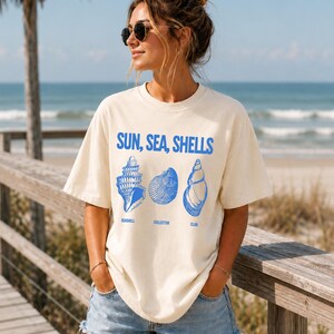 May include: An off-white short-sleeve t-shirt featuring blue graphic text that reads SUN, SEA, SHELLS and SEASHELL COLLECTOR CLUB, accompanied by illustrations of three distinct seashells in a matching blue ink color.
