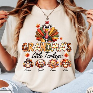 May include: An ivory t-shirt featuring a large graphic of a turkey with colorful plaid feathers above the text Grandma's Little Turkeys. Below the text are four smaller cartoon turkeys with the names Sophia, David, Emma, and Alex printed underneath.