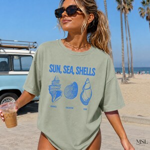 K&ouml;nnte beinhalten: Ein salbeigr&uuml;nes Kurzarm-T-Shirt mit dem blauen Schriftzug "SUN, SEA, SHELLS" &uuml;ber drei detaillierten blauen Illustrationen verschiedener Muscheln. Unter den Muscheln steht der Text "SEASHELL", "COLLECTOR" und "CLUB" in blauer Schrift.
