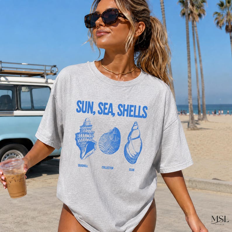 K&ouml;nnte beinhalten: Ein hellgraues Kurzarm-T-Shirt mit dem blauen Schriftzug SUN, SEA, SHELLS &uuml;ber drei blauen Strichzeichnungen von Muscheln. Unter den Muscheln stehen die W&ouml;rter SEASHELL, COLLECTOR und CLUB in blauen Gro&szlig;buchstaben.