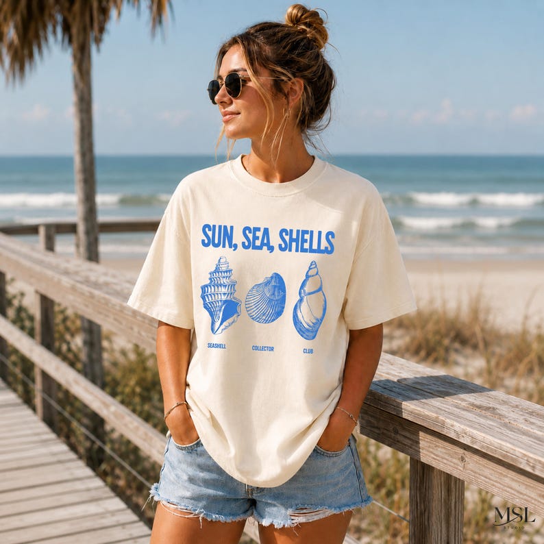 May include: An off-white short-sleeve t-shirt featuring blue graphic text that reads SUN, SEA, SHELLS and SEASHELL COLLECTOR CLUB, accompanied by illustrations of three distinct seashells in a matching blue ink color.