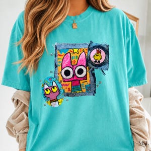 May include: A turquoise t-shirt featuring a collage of colorful, hand-drawn style illustrations. The design includes a large pink cat face, a smaller purple cat, and a yellow bird, all accented with safety pins and frayed fabric edges.