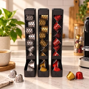 Peut inclure: Trois distributeurs de capsules de café noirs verticaux avec un panneau avant à motif en nid d'abeille. Chaque distributeur contient une colonne de capsules de café argentées, dorées et rouges. La base porte un logo N et un plateau ouvert.