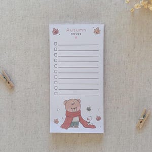 May include: A rectangular notepad titled Autumn notes with a checklist of ten lines. The design features a brown bear wearing a long red scarf, a small white bird, and scattered autumn leaves in shades of orange and brown on a light background.