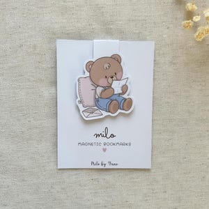 Puede incluir: Un marcapáginas magnético con la ilustración de un oso marrón con peto azul, sentado contra una almohada rosa claro mientras sostiene una carta blanca. El marcapáginas está sujeto a una tarjeta blanca con el texto "milo MAGNETIC BOOKMARKS" y "milo by Vane" impreso en negro, junto a un pequeño icono de corazón rosa.
