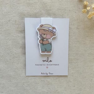 Puede incluir: Un marcapáginas magnético con la ilustración de un oso marrón que lleva un sombrero beige con una cinta morada clara y un peto azul, sosteniendo un pequeño ramo de lavanda. El marcapáginas está sujeto a una tarjeta blanca con "milo MAGNETIC BOOKMARKS".