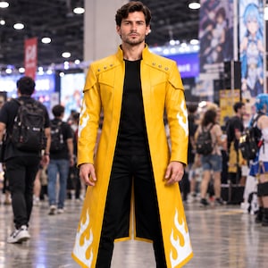 Puede incluir: Un abrigo largo de color amarillo brillante con cuello alto, hombros estructurados y patrones blancos en forma de llama a lo largo de las mangas y el dobladillo. El abrigo se lleva sobre una camiseta negra lisa y pantalones negros, con botas de cuero negro.