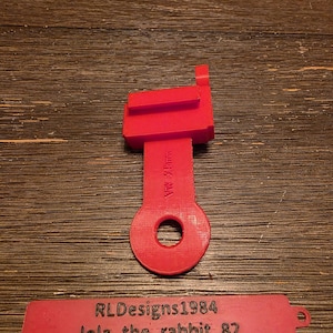 May include: A bright red, 3D-printed plastic part with a rectangular top, a stem with text, and a circular hole at the bottom. A separate red tag displays the text "RLDesigns1984" and "lola_the_rabbit_82".
