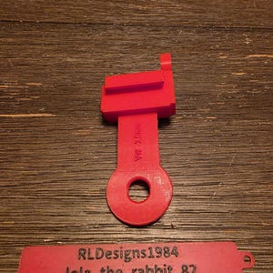 May include: A bright red, 3D-printed plastic part with a rectangular top, a stem with text, and a circular hole at the bottom. A separate red tag displays the text "RLDesigns1984" and "lola_the_rabbit_82".