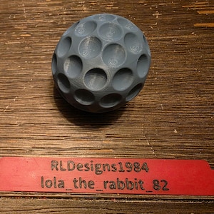 May include: A gray, spherical object with numerous circular indentations, resembling a golf ball, rests on a wooden surface. A red tag below it displays the text "RLDesigns1984 lola_the_rabbit_82".