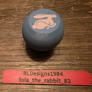 May include: A blue, spherical object with a pink rabbit design on top. Below it is a red tag with the text "RLDesigns1984 lola_the_rabbit_82" engraved on it. The items are on a wooden surface.