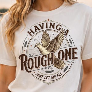 Op de afbeelding: Een off-white t-shirt met een cirkelvormige afbeelding van een vliegende duif in het midden. De tekst "HAVING A ROUGH ONE" staat in grote bruine letters, met "JUST LET ME FLY" eronder. Het ontwerp bevat kleine sterren, veren en lijnen.
