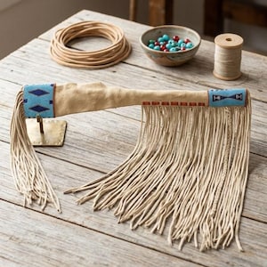 May include: A tan leather object with long fringe, featuring two sections of blue, white, and dark blue geometric beadwork at the ends and a row of small red rectangular beads along the center. The item is displayed on a small metal stand.
