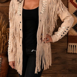 May include: A beige suede jacket featuring long fringe detailing along the chest, shoulders, and sleeves. The garment includes silver-toned metal studs and white leather lacing accents along the front placket and down the length of the sleeves.