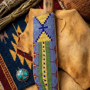 May include: A beaded knife sheath with a wooden handle, featuring a geometric design in blue, yellow, and green beads. The sheath rests on a tan leather surface, with a turquoise and silver button and woven textile in the background.