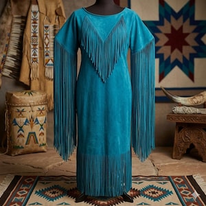 May include: A turquoise suede dress with long sleeves and fringe detailing. The dress features fringe along the neckline, sleeves, and hem. The dress is displayed on a mannequin, showcasing its design and texture. The dress is part of a collection of Native American-inspired clothing.