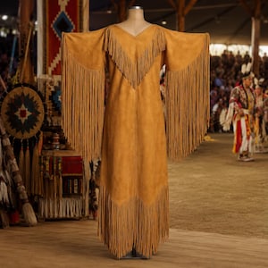 May include: A tan suede dress with long sleeves and a V-neckline. The dress features long fringe detailing along the sleeves, neckline, and hem. The dress is displayed on a mannequin in a well-lit indoor setting, with a crowd in the background.